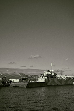 A large industrial ship docked beside a concrete barrier in a harbor setting. The ship features a smokestack and appears to have a camouflaged pattern on its hull. Surrounding the dock are other industrial structures and equipment, partially visible hills, and a partially cloudy sky overhead.
