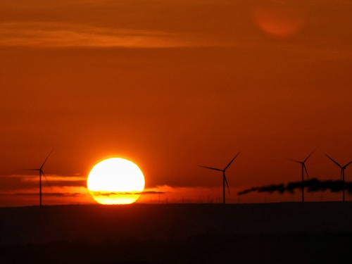 A beautiful sunset with a large, glowing sun near the horizon, casting an orange hue across the sky. In the foreground, several wind turbines are silhouetted against the vibrant backdrop, suggesting a sustainable energy scene.