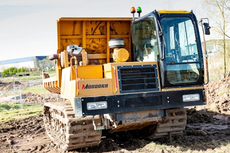 A large yellow Morooka tracked vehicle is positioned on a muddy construction site. It features a spacious cabin with large windows and is equipped with lights on top. There is earth and construction fencing in the background, indicating an active construction area.