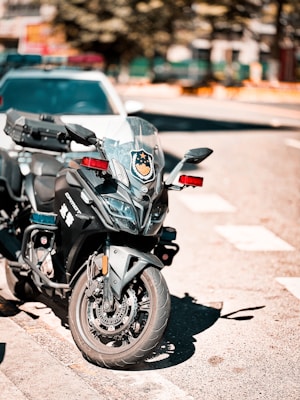 A black police motorcycle with red and blue emergency lights is parked on a city street beside a white vehicle. The motorcycle has a shield emblem on the windshield, and the pavement around it is sunlit.