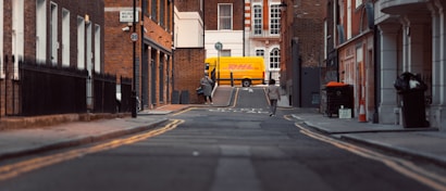 A narrow urban street scene with brick buildings on either side. In the background, a bright yellow DHL delivery van is parked. Two pedestrians are walking along the street, and there are several street signs and road markings visible. The atmosphere is calm, with a focus on the architecture and the delivery van.