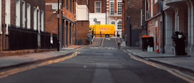 A narrow urban street scene with brick buildings on either side. In the background, a bright yellow DHL delivery van is parked. Two pedestrians are walking along the street, and there are several street signs and road markings visible. The atmosphere is calm, with a focus on the architecture and the delivery van.