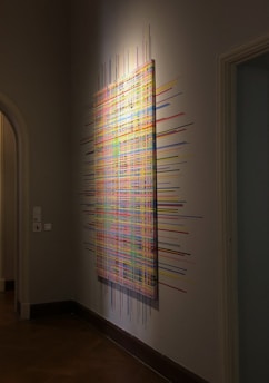 An abstract artwork is mounted on a wall in what appears to be a gallery space. The piece consists of a complex, multi-colored grid with lines extending beyond the canvas edges, creating a sense of movement and expansion. The surrounding area is dimly lit, highlighting the vibrant colors of the art.