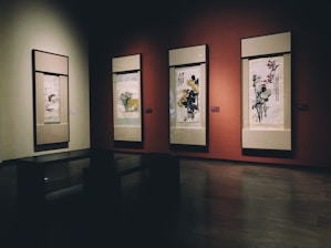 A dimly lit gallery room showcasing four traditional Asian scroll paintings, each framed and displayed on a deep red wall. The scrolls depict various floral motifs and include calligraphy. The paintings are subtly illuminated by soft lighting, enhancing the rich colors. A dark-colored bench is placed in the foreground, facing the artworks.