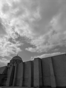A large, imposing stone building features a prominent dome and towering walls with vertical supports. The sky above is filled with a vast expanse of clouds, giving a dramatic effect. The architecture appears historic and robust, emphasizing strength and stability.
