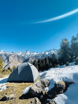A gray tent set up on a grassy area surrounded by snow with a backdrop of towering, snow-capped mountains under a clear blue sky. Pine trees are visible in the midground, adding to the natural scenery.