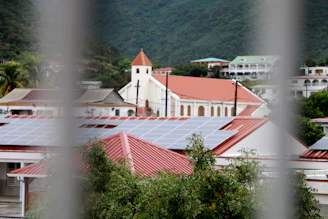 A view of buildings with red roofs and solar panels, set against a backdrop of lush green hills. In the middle ground, a church with a steeple stands out. The foreground is partially obscured by metal bars and leafy vegetation.