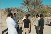 A group of men are gathered in an outdoor setting with palm trees and a stone wall in the background. They appear to be engaged in a discussion. One man is wearing a mask, while others are dressed in various casual and traditional attire. A motorcycle is parked nearby.