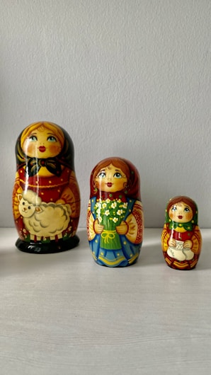 Three Russian nesting dolls, each with a unique design and size, arranged side by side on a light surface against a plain background. The largest doll features a sheep design, while the smaller ones depict floral and traditional motifs.