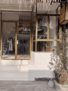 A boutique storefront with glass windows and doors, featuring a wooden frame. Inside, clothing items are displayed on racks, and a woven lampshade hangs from the ceiling. A potted plant is placed near the entrance, adding a touch of greenery. The exterior design includes a thatched awning.