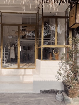 A boutique storefront with glass windows and doors, featuring a wooden frame. Inside, clothing items are displayed on racks, and a woven lampshade hangs from the ceiling. A potted plant is placed near the entrance, adding a touch of greenery. The exterior design includes a thatched awning.