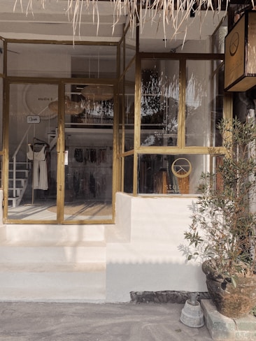 A boutique storefront with glass windows and doors, featuring a wooden frame. Inside, clothing items are displayed on racks, and a woven lampshade hangs from the ceiling. A potted plant is placed near the entrance, adding a touch of greenery. The exterior design includes a thatched awning.
