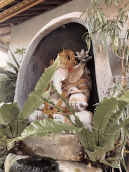 A stone statue of Ganesha adorned with intricate golden accessories is situated within a small alcove surrounded by tropical plants. The statue is detailed with a serene expression and upraised hand gestures symbolizing blessings and protection.