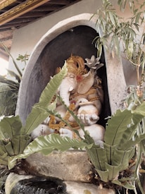 A stone statue of Ganesha adorned with intricate golden accessories is situated within a small alcove surrounded by tropical plants. The statue is detailed with a serene expression and upraised hand gestures symbolizing blessings and protection.