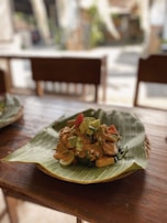 A dish arranged on a banana leaf placed over a wooden table. The meal consists of vegetables and pieces of tofu or tempeh topped with a rich-looking sauce. The setting appears to be a casual dining area with wooden chairs in the background.