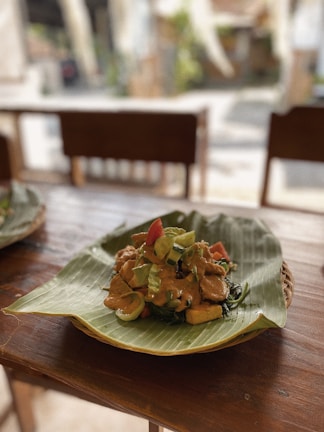 A dish arranged on a banana leaf placed over a wooden table. The meal consists of vegetables and pieces of tofu or tempeh topped with a rich-looking sauce. The setting appears to be a casual dining area with wooden chairs in the background.