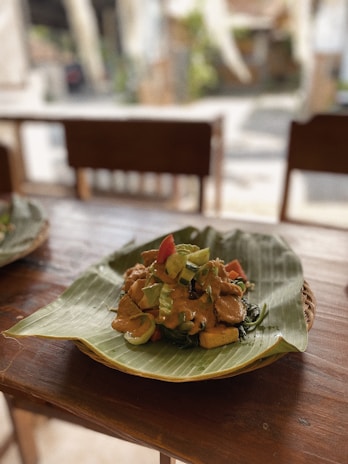 A dish arranged on a banana leaf placed over a wooden table. The meal consists of vegetables and pieces of tofu or tempeh topped with a rich-looking sauce. The setting appears to be a casual dining area with wooden chairs in the background.