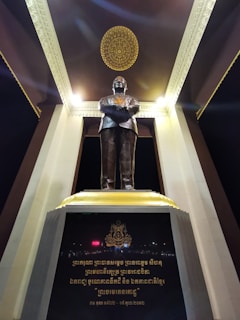 A bronze statue of a man stands with folded arms on a pedestal, situated under an ornately decorated ceiling with intricate golden patterns. The structure is framed by large columns, and inscriptions in another language appear on the base of the statue.