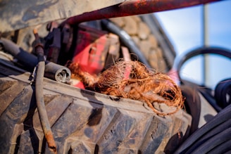 A large, rugged tire with a metal rod and some wiring partially covered by a bunch of frayed brown fibers. The background is slightly blurred, featuring elements in various shades, with a glimpse of red equipment.