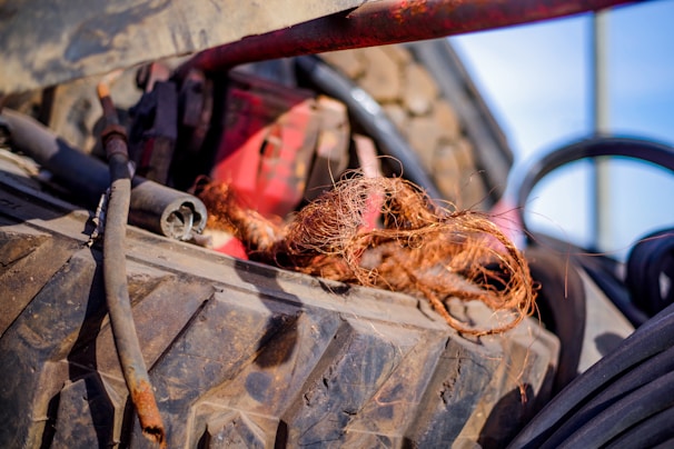 A large, rugged tire with a metal rod and some wiring partially covered by a bunch of frayed brown fibers. The background is slightly blurred, featuring elements in various shades, with a glimpse of red equipment.