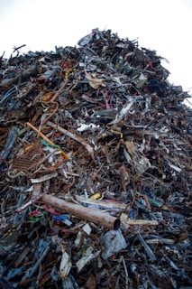 A large heap of assorted scrap metal and debris, including rusted pipes, wires, metal sheets, and various other discarded items, forming a significant pile. The background is a clear sky.