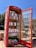 A red British-style telephone booth repurposed as a small library, with shelves filled with a variety of books. The booth is labeled as a 'BOOK BOX' at the top. The surrounding area is outdoors on a clear, sunny day, with trees and a stone wall visible in the background.