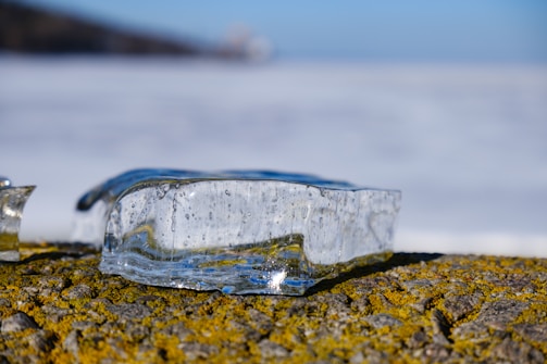 A clear, rectangular block of ice rests on a moss-covered stone surface. The ice contains small bubbles and reflects light, creating a shimmering effect. The background is blurred, suggesting an outdoor setting with a distant horizon.