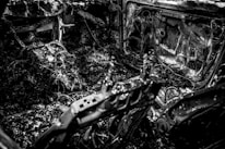 A charred and burnt-out interior of a vehicle, displaying twisted metal, exposed wires, and debris scattered throughout. The surfaces are covered in soot and ash, suggesting severe fire damage.