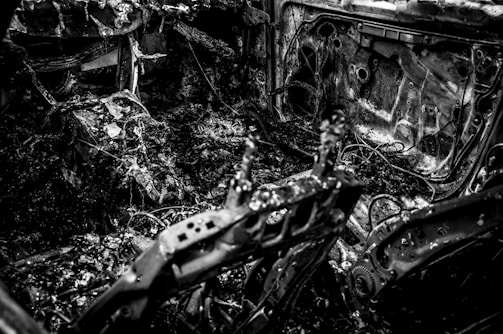 A charred and burnt-out interior of a vehicle, displaying twisted metal, exposed wires, and debris scattered throughout. The surfaces are covered in soot and ash, suggesting severe fire damage.