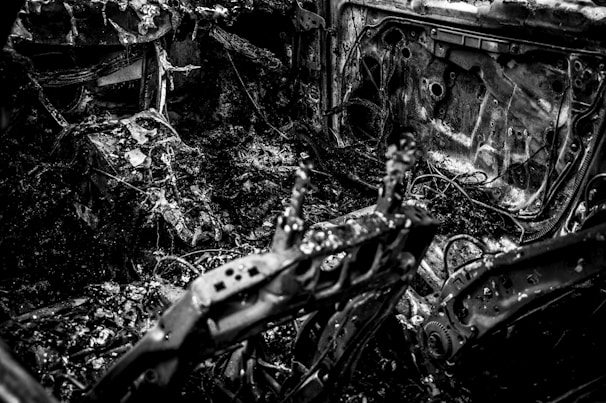 A charred and burnt-out interior of a vehicle, displaying twisted metal, exposed wires, and debris scattered throughout. The surfaces are covered in soot and ash, suggesting severe fire damage.