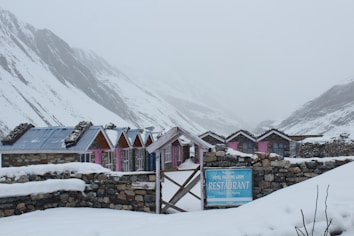 A row of stone cottages with colorful wooden doors and metal roofs stretches across a snowy landscape. The mountainous backdrop is shrouded in mist, adding to the serene and chilly atmosphere. A rustic signboard in front advertises a restaurant within the establishment.