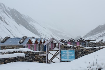 A row of stone cottages with colorful wooden doors and metal roofs stretches across a snowy landscape. The mountainous backdrop is shrouded in mist, adding to the serene and chilly atmosphere. A rustic signboard in front advertises a restaurant within the establishment.