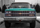 A vintage Chevrolet truck is parked on a cobblestone street, centered in the image. It has a teal body with chrome accents and a distinct front grille featuring the Chevrolet logo. The truck's headlights are symmetrical and operational. There are potted plants and modern buildings in the background, adding an urban setting.