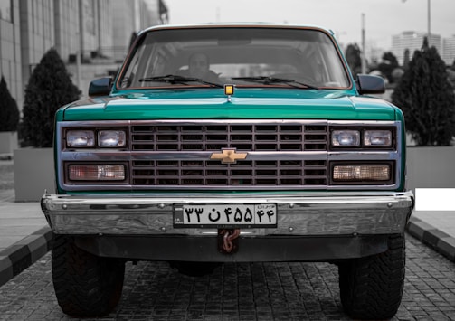 A vintage Chevrolet truck is parked on a cobblestone street, centered in the image. It has a teal body with chrome accents and a distinct front grille featuring the Chevrolet logo. The truck's headlights are symmetrical and operational. There are potted plants and modern buildings in the background, adding an urban setting.