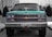 A vintage Chevrolet truck is parked on a cobblestone street, centered in the image. It has a teal body with chrome accents and a distinct front grille featuring the Chevrolet logo. The truck's headlights are symmetrical and operational. There are potted plants and modern buildings in the background, adding an urban setting.