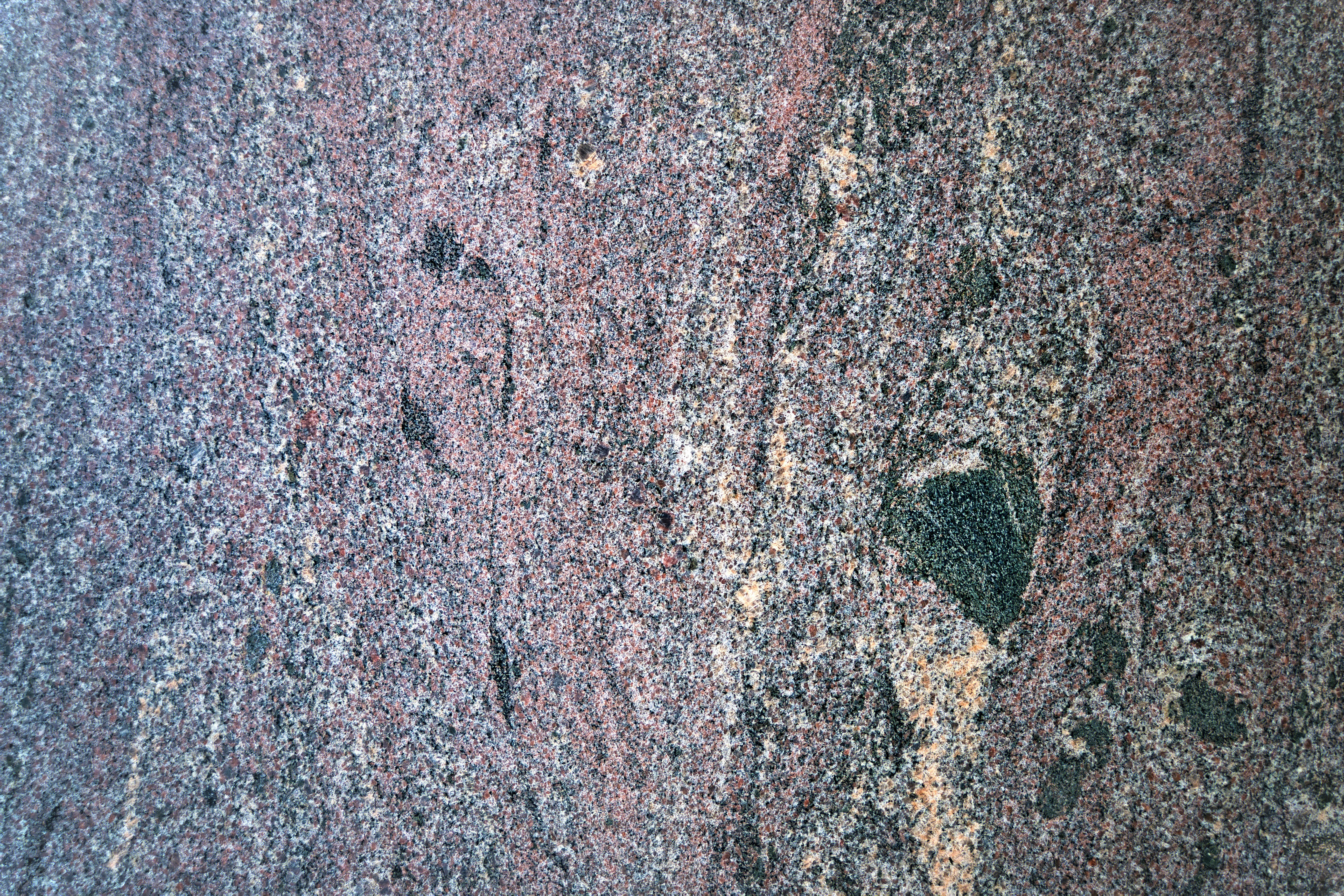 A close-up view of a rough, multicolored granite surface with a speckled and slightly wavy pattern. The dominant colors include shades of pink, gray, black, and some tan areas. The texture appears coarse and varied.