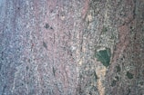 A close-up view of a rough, multicolored granite surface with a speckled and slightly wavy pattern. The dominant colors include shades of pink, gray, black, and some tan areas. The texture appears coarse and varied.