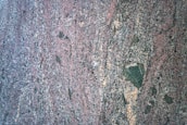 A close-up view of a rough, multicolored granite surface with a speckled and slightly wavy pattern. The dominant colors include shades of pink, gray, black, and some tan areas. The texture appears coarse and varied.
