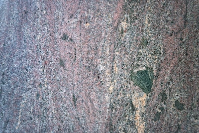 A close-up view of a rough, multicolored granite surface with a speckled and slightly wavy pattern. The dominant colors include shades of pink, gray, black, and some tan areas. The texture appears coarse and varied.