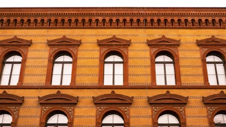 A brick building facade with arched windows and intricate patterns. The structure features a warm color palette, with decorative brickwork and detailed cornices. The windows are symmetrically arranged across two floors.