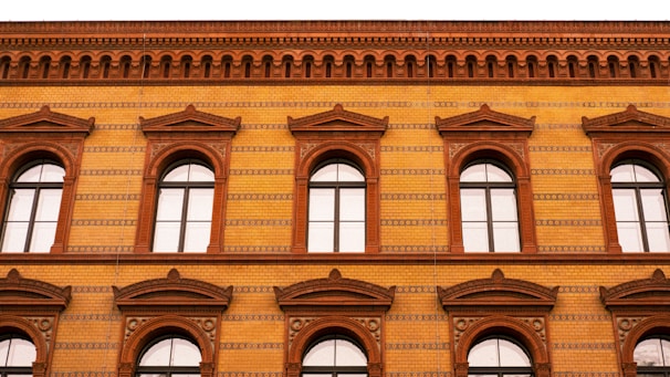 A brick building facade with arched windows and intricate patterns. The structure features a warm color palette, with decorative brickwork and detailed cornices. The windows are symmetrically arranged across two floors.