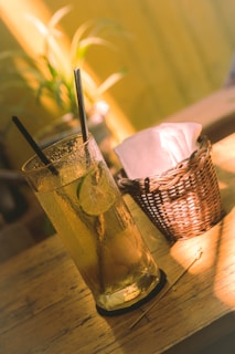 A tall glass filled with an iced drink, garnished with lime slices, sits on a wooden table. Two black straws are immersed in the drink. Next to the glass, a wicker basket holds white napkins. A plant with green leaves is blurred in the background, suggesting a natural, relaxed setting.