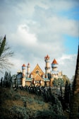A grand and majestic castle with multiple towers and spires sits atop a lush green hill surrounded by tall trees. The castle’s architecture features a mix of medieval and fantasy elements, with pointed turrets and ornate detailing. The sky is partly cloudy, creating a dramatic backdrop.