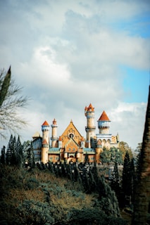 A grand and majestic castle with multiple towers and spires sits atop a lush green hill surrounded by tall trees. The castle’s architecture features a mix of medieval and fantasy elements, with pointed turrets and ornate detailing. The sky is partly cloudy, creating a dramatic backdrop.
