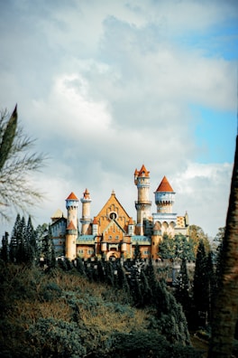 A grand and majestic castle with multiple towers and spires sits atop a lush green hill surrounded by tall trees. The castle’s architecture features a mix of medieval and fantasy elements, with pointed turrets and ornate detailing. The sky is partly cloudy, creating a dramatic backdrop.