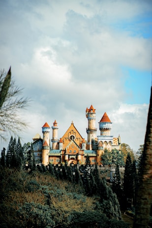 A grand and majestic castle with multiple towers and spires sits atop a lush green hill surrounded by tall trees. The castle’s architecture features a mix of medieval and fantasy elements, with pointed turrets and ornate detailing. The sky is partly cloudy, creating a dramatic backdrop.