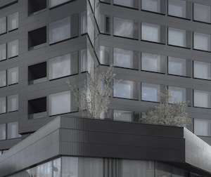 A modern architectural facade with a grid pattern of reflective windows. The building features dark-colored cladding and a minimalist design, with some greenery visible on a lower rooftop. The structure conveys a sleek and contemporary aesthetic.