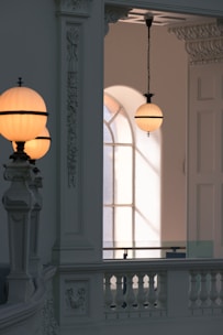 A classic interior with ornate architectural details, featuring spherical pendant lamps with a soft glow. Light filters through a tall arched window, casting shadows on the wall and highlighting the decorative moldings and the balustrade.