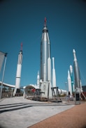 A collection of large, metallic rockets standing upright against a clear blue sky, with some red and black detailing. The area appears to be an outdoor exhibition, with visitors walking around and observing the display. There is a concrete path and platforms surrounding the rockets, and some greenery can be seen in the background.