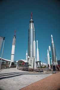 A collection of large, metallic rockets standing upright against a clear blue sky, with some red and black detailing. The area appears to be an outdoor exhibition, with visitors walking around and observing the display. There is a concrete path and platforms surrounding the rockets, and some greenery can be seen in the background.